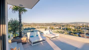 A rooftop terrace with two white lounge chairs, a palm tree, and a city view.