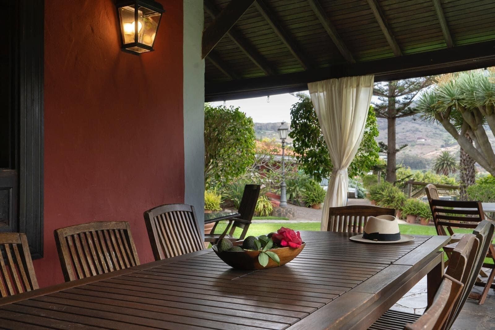 A wooden table with chairs under a covered patio, a bowl of fruit, and a hat on the table.