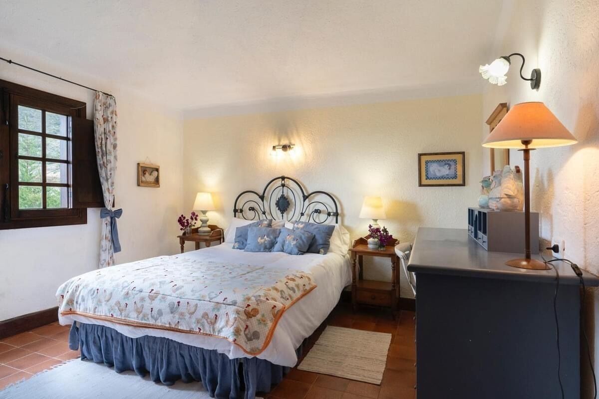 Un dormitorio con cama, dos mesitas de noche con lámparas, un aparador y una ventana con cortinas.