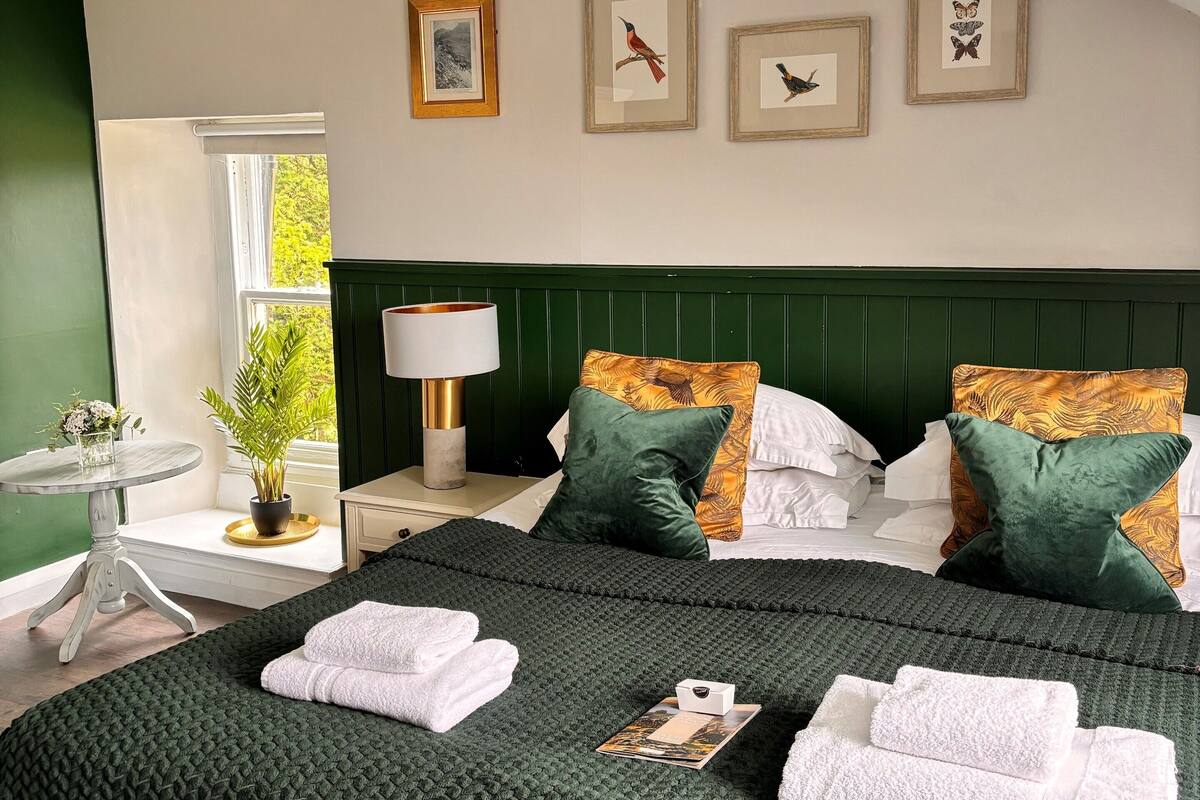 A neatly made bed with green and white pillows, a bedside table with a lamp, and framed artwork on the wall.