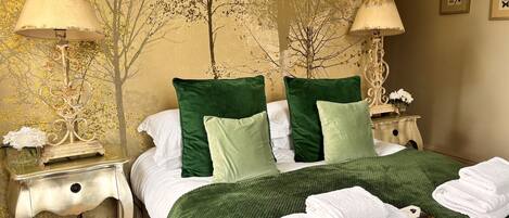 Ein ordentlich gemachtes Bett mit grünen und weißen Kissen, einer grünen Decke und weißen Handtüchern auf einem strukturierten grünen Teppich. Es gibt zwei Nachttische, jeweils mit einer Lampe und einer kleinen Dekoration.