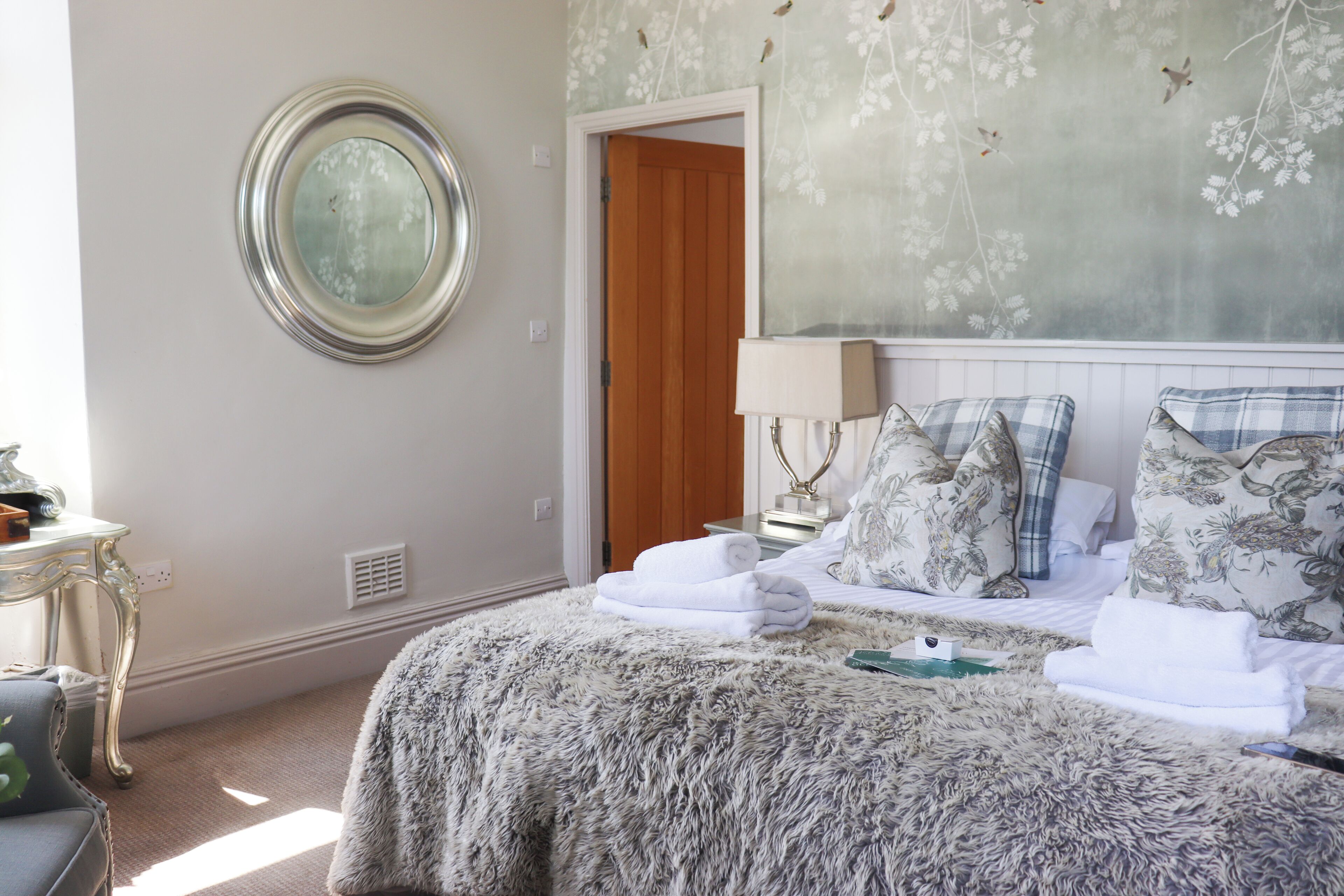 Una cama bien hecha con un edredón esponjoso, un espejo redondo en la pared y un papel tapiz con motivos florales.