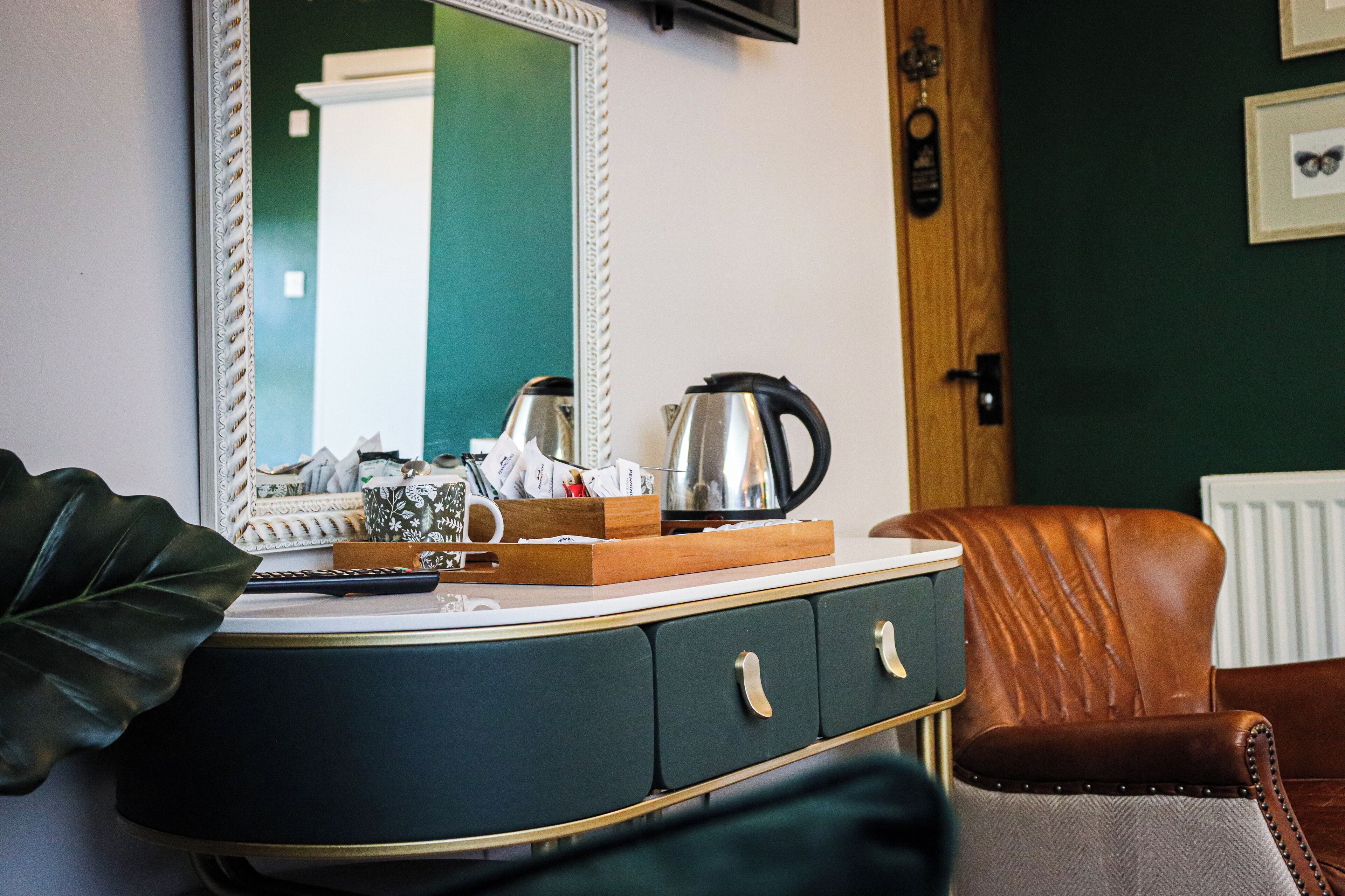 A room with a dark green wall, a white-framed mirror, a wooden cabinet with drawers, a brown leather chair, and a radiator.