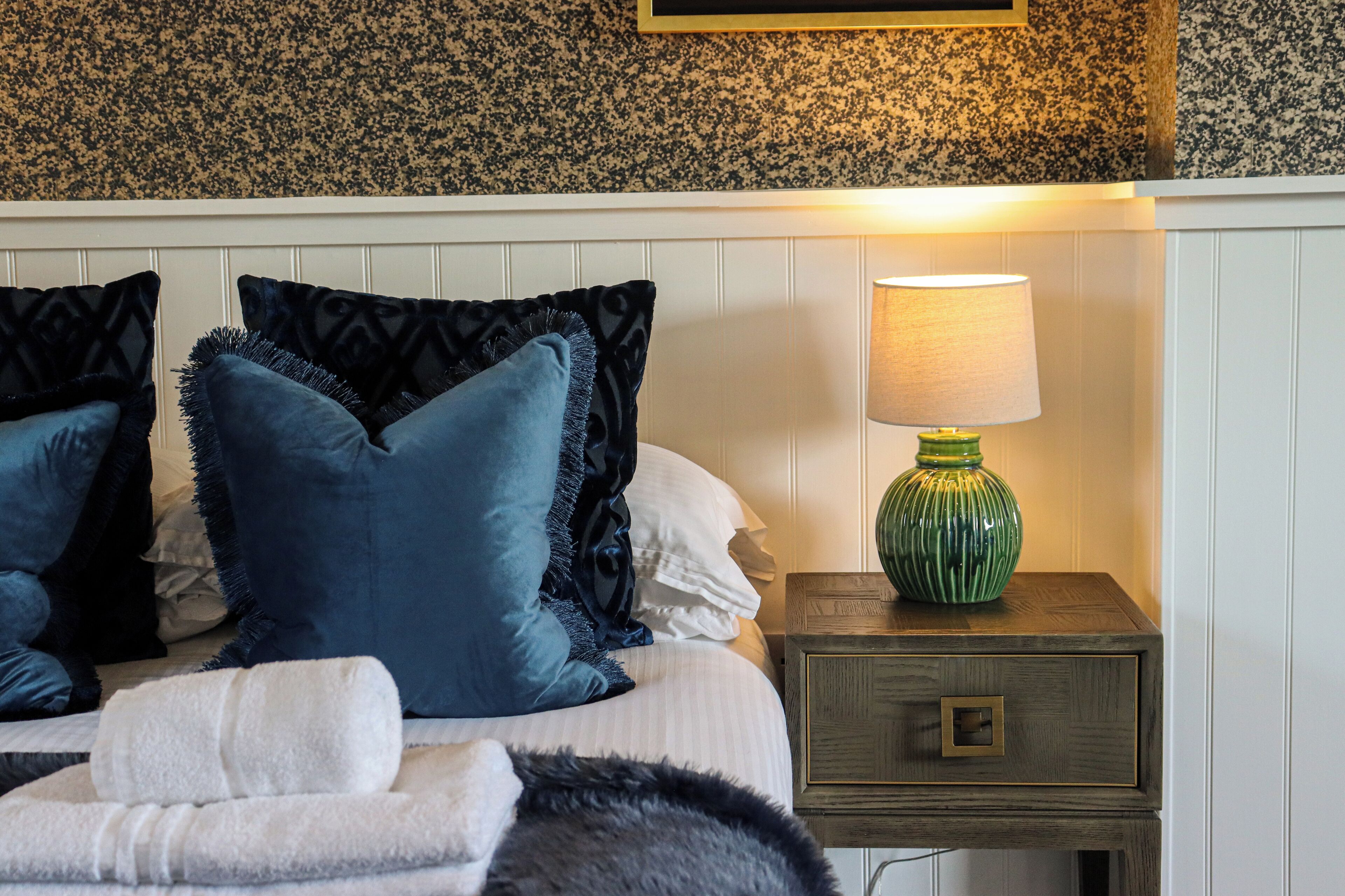 A neatly made bed with blue and white pillows, a green lamp on a wooden nightstand, and a patterned wall behind.