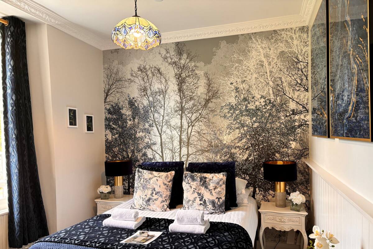 A bedroom with a bed, two bedside tables, a chandelier, and a mural of trees on the wall.