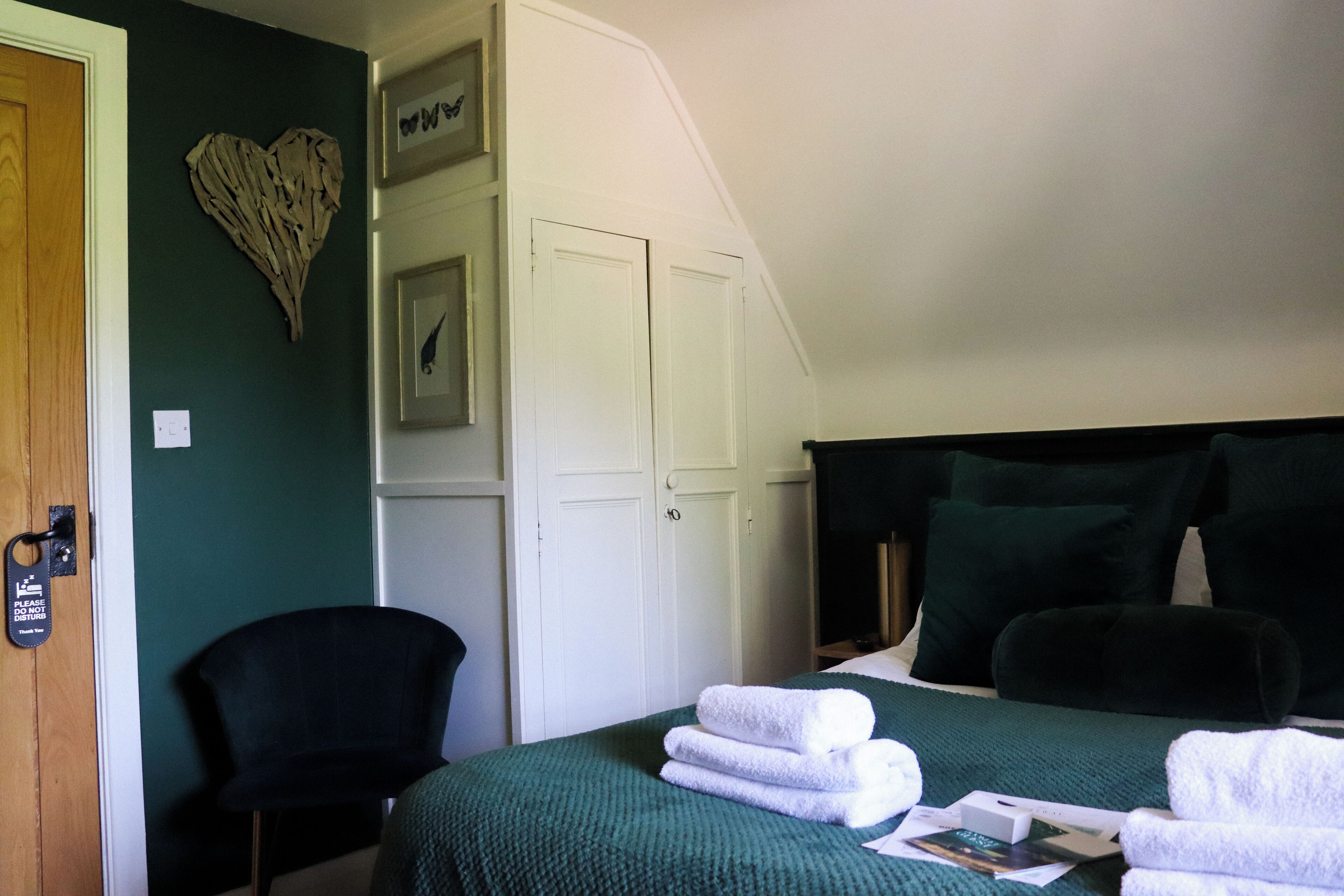 Un dormitorio con cama, ropa de cama verde, una silla, un armario y una decoración con forma de corazón en la pared.