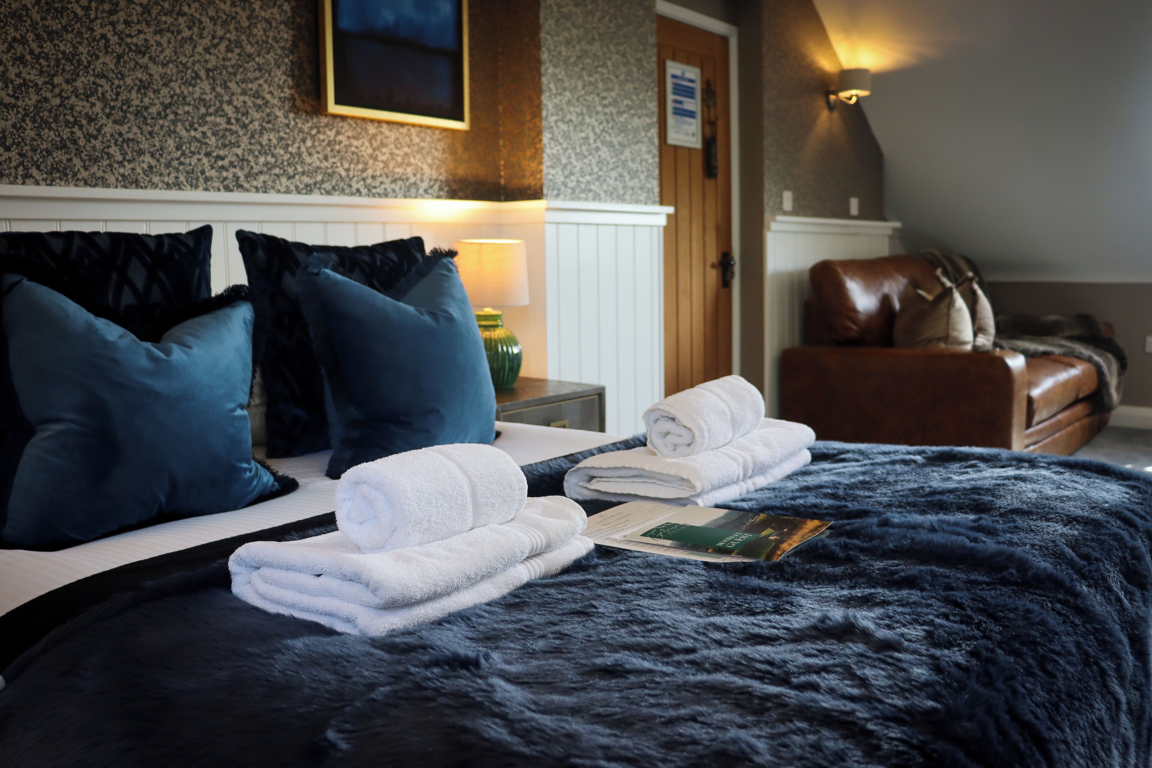 A neatly made bed with a dark blue blanket, white towels, and a folded magazine on a hotel room with a wooden headboard, a brown leather chair, and a wall-mounted lamp.