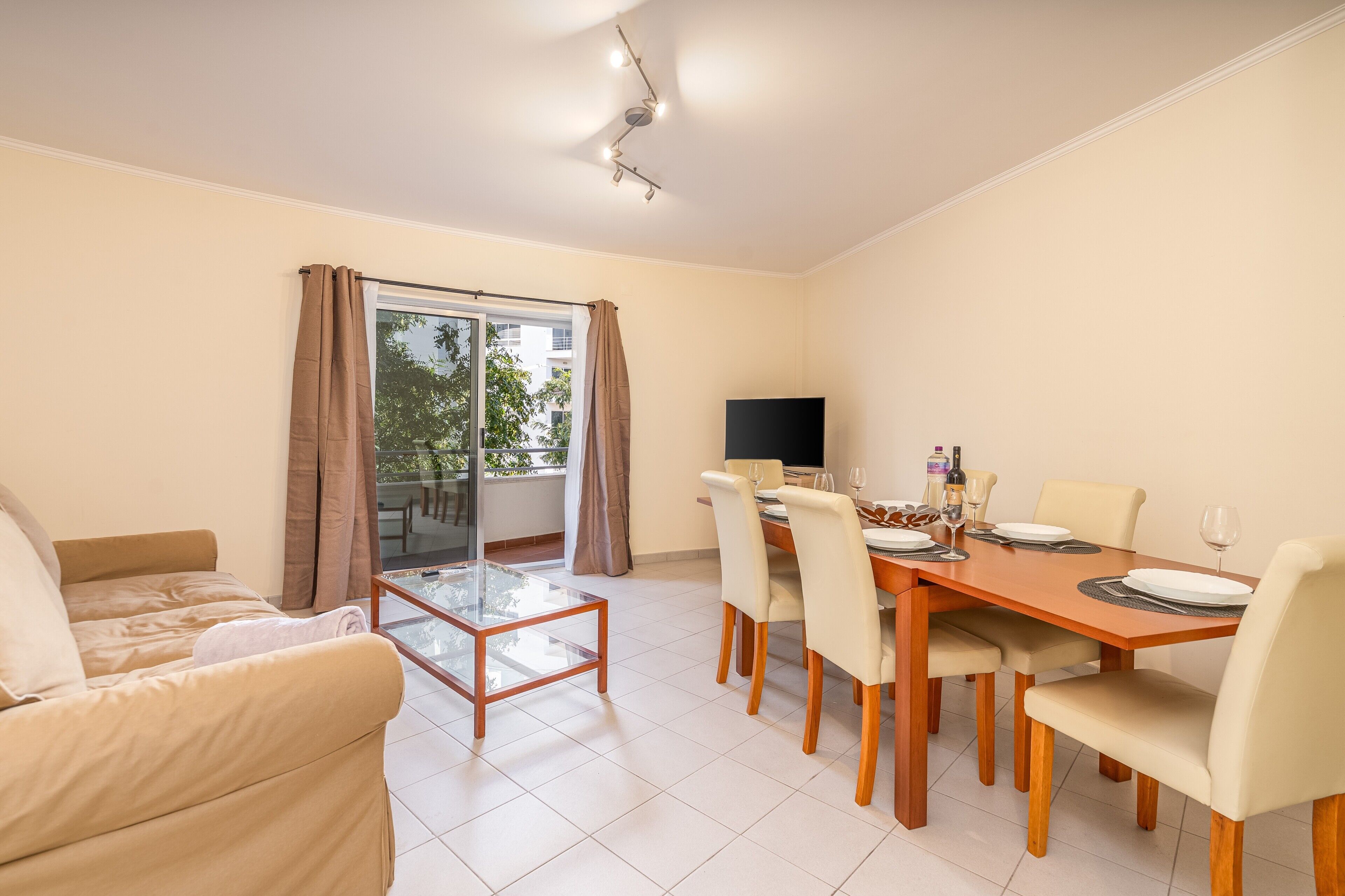 Uma sala moderna com sofá, mesa de centro de vidro, mesa de jantar com cadeiras, televisão de tela plana e porta de vidro deslizante que dá para uma área externa.