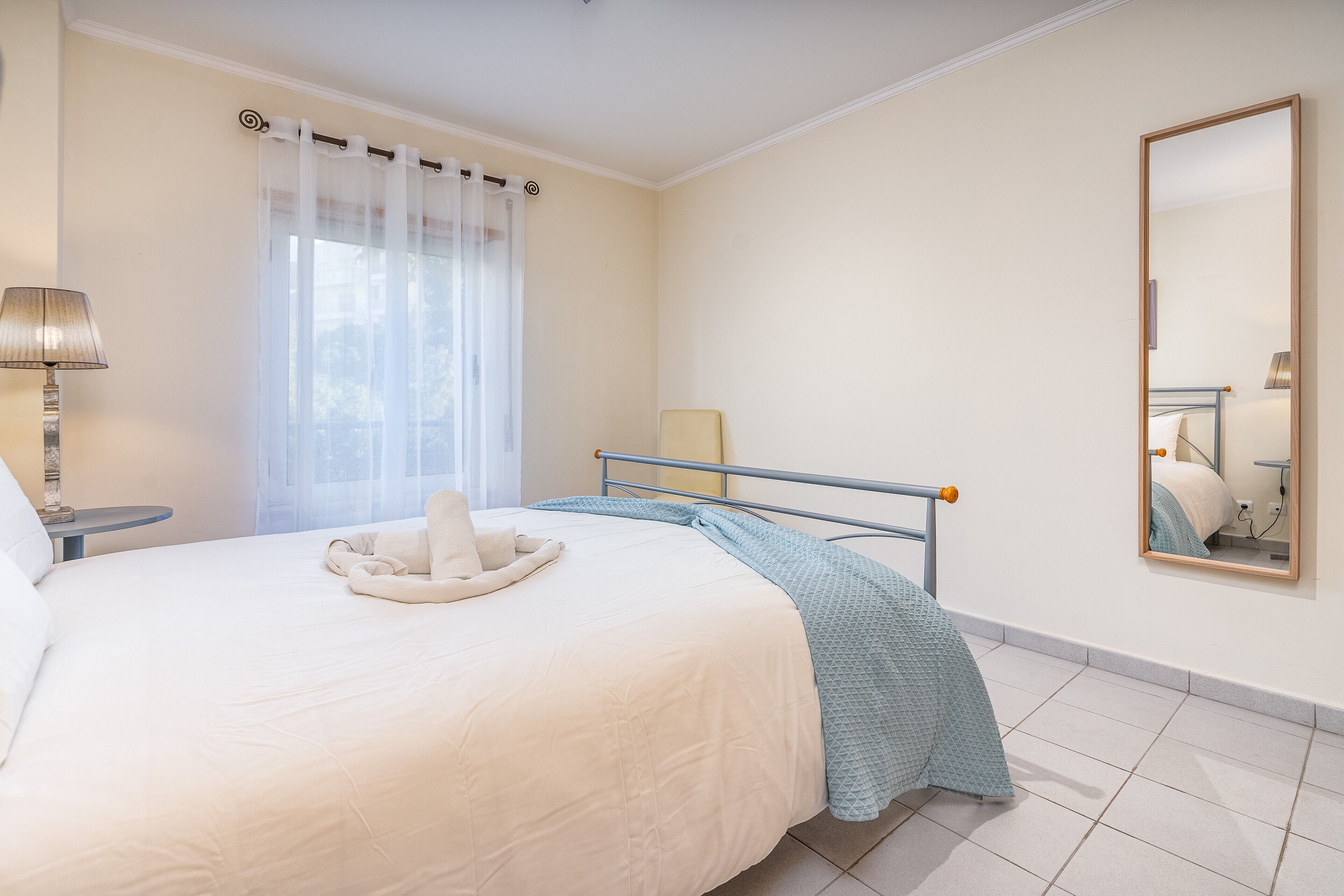 Une chambre d’hôtel avec un lit, une table de chevet, une lampe, une fenêtre avec des rideaux légers et un miroir au mur.