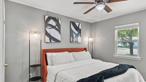 A bedroom with a bed, a ceiling fan, two wall-mounted floor lamps, and a window with a view of a tree and house. - CASA Noa Noa (Euless)