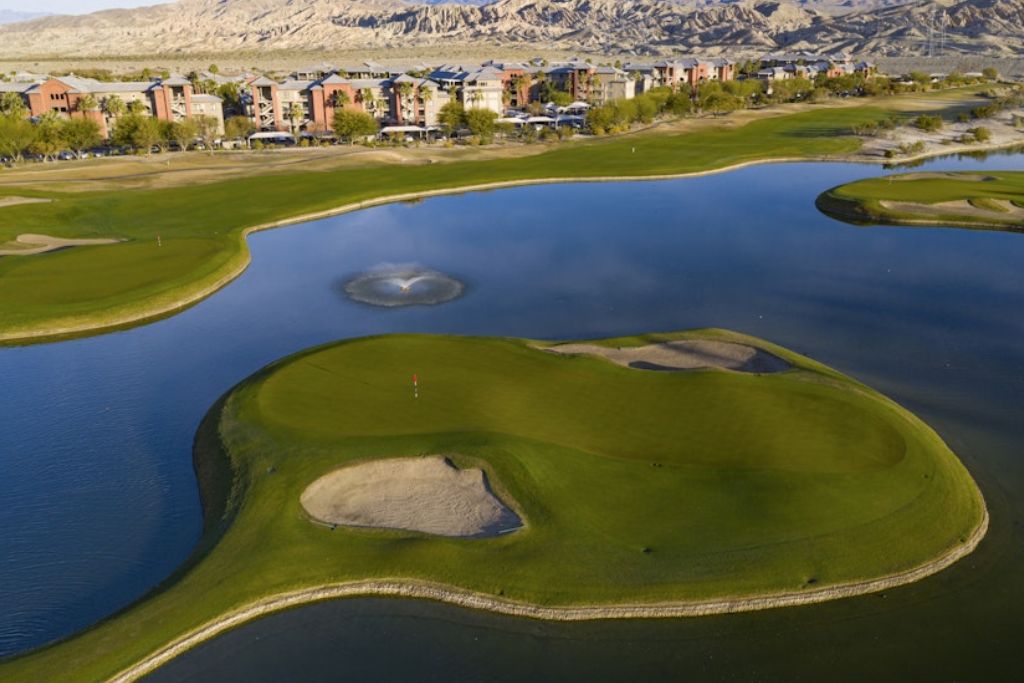 Un campo de golf con un estanque grande, búnkeres de arena y edificios al fondo.