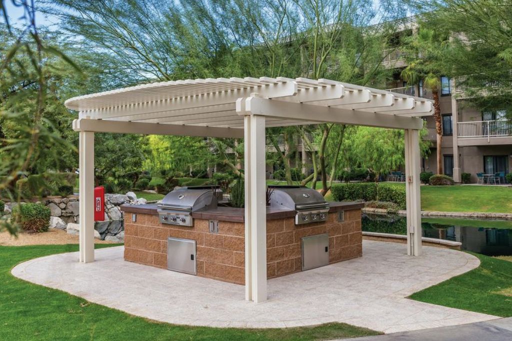A covered outdoor grill area with a pergola, surrounded by greenery and a building in the background.