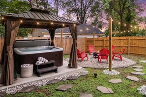 A hot tub with a canopy, surrounded by red chairs and a fire pit, set in a backyard with a wooden fence and string lights.