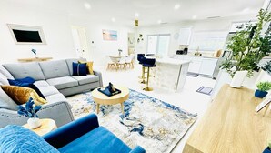 Een moderne woonkamer met een grijze bank, blauwe fauteuils en een salontafel. Achtergrond is een keuken met witte kasten en een eettafel.