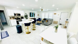Een moderne keuken met witte kasten, een centraal eiland en barstoelen. Er is een zithoek met een witte tafel en stoelen, een grijze bank en een klein vloerkleed.