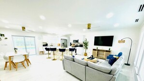 Een moderne woonkamer met een grijze bank, een eettafel met witte stoelen, een keuken met witte kasten en een flatscreen aan de muur.