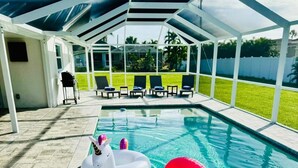 Een zwembad met drijvende zwanen en eenhoorn-speelgoed, een overdekt terras met lounge stoelen, en uitzicht op een grasveld.