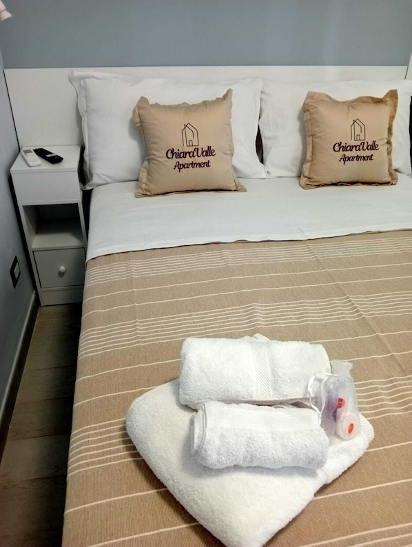 A hotel room with a bed, two pillows with the hotel name, a nightstand with a remote, and a folded towel on the bed.