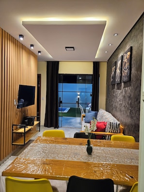 A modern living room with a flat-screen TV, a sofa, a dining table with yellow chairs, and a large window offering a view of the outdoors. - Casa Hana (Camaçari)