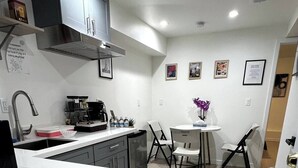 Una cocina moderna con una isla central, una zona de comedor con mesa y sillas blancas, y un pequeño rincón de estar con un jarrón de flores morado.