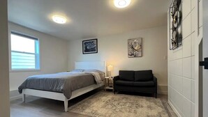 Un dormitorio con cama, mesita de noche, sofá negro, alfombra y una obra de arte colgada en la pared.