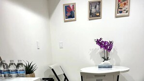 Cocina moderna con lavavajillas integrado, encimera blanca y mesa redonda con dos sillas. En la pared hay obras de arte enmarcadas y una planta en maceta sobre la encimera.