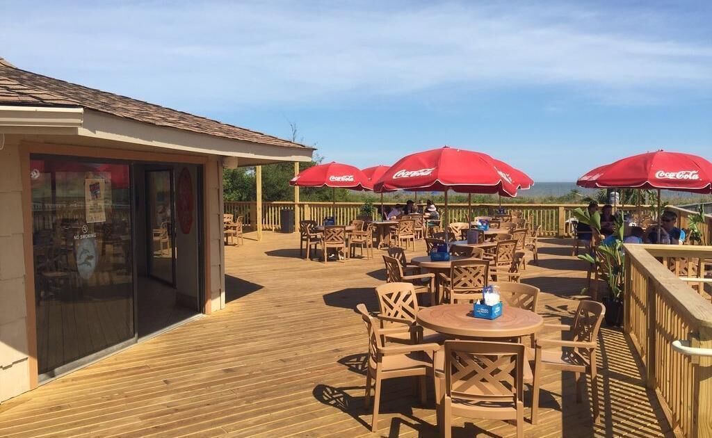 An outdoor patio with wooden tables and chairs, red umbrellas, and a view of the ocean.