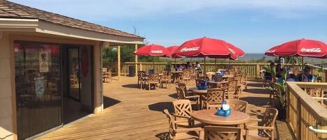 An outdoor patio with wooden tables and chairs, red umbrellas, and a view of the ocean.