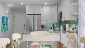 A modern kitchen with white cabinets, a stainless steel refrigerator, and a dining area with a glass table and white chairs.