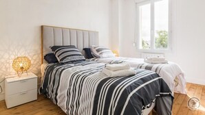 Un dormitorio moderno con dos camas, ropa de cama a rayas, una mesita de noche con lámpara y una ventana con vistas.