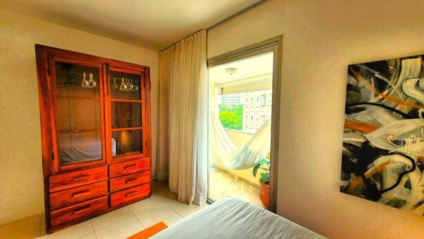 A bedroom with a wooden wardrobe, a bed, a large abstract painting, and a view of greenery through a window. - Floripa, small and sexy. (Centro Florianópolis)