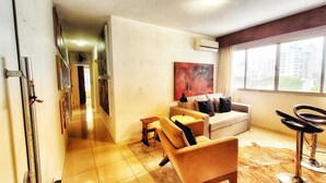 A modern living room with a sofa, a small table, and a bar stool. There is a painting on the wall and an air conditioning unit mounted above the sofa. - Floripa, small and sexy. (Centro Florianópolis)
