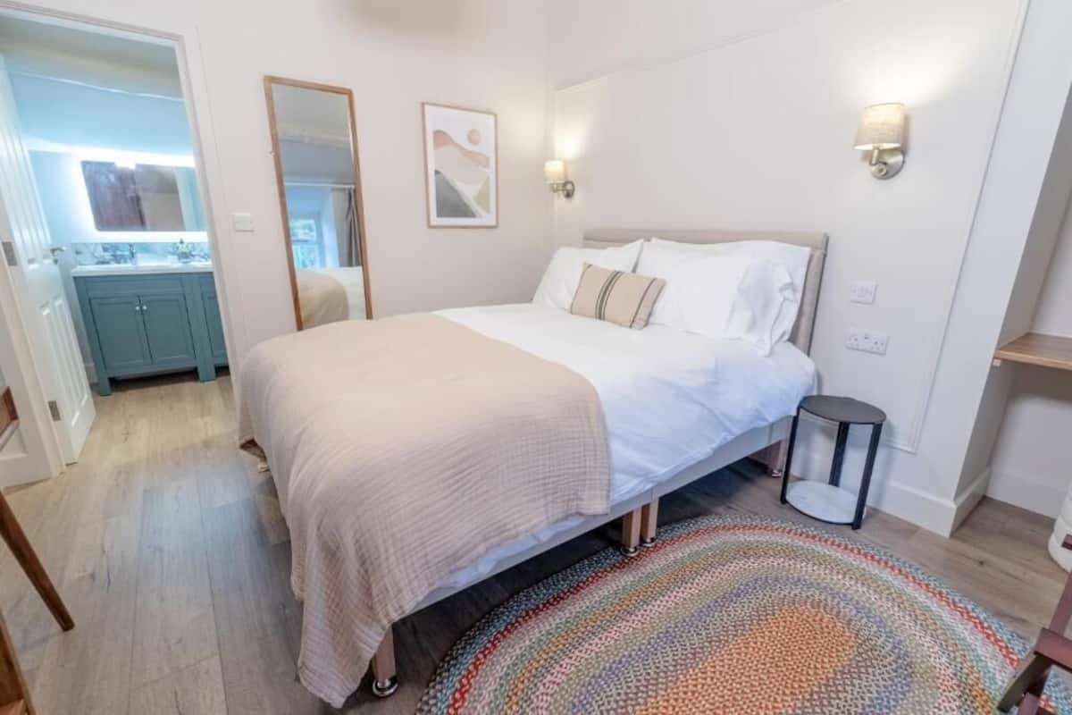 A bedroom with a bed, bedside table, wall-mounted light, and a door leading to a bathroom.