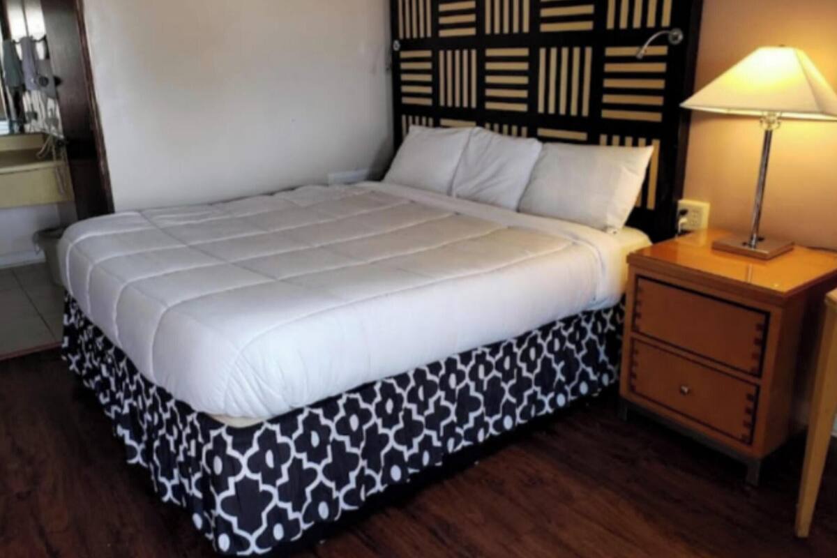 Una cama bien hecha con falda estampada, una lámpara de noche y una mesita de noche de madera con cajones.