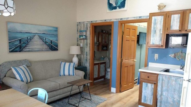 A living room with a sofa, a coffee table, and a painting of a pier. There is a kitchen with wooden cabinets and a microwave.