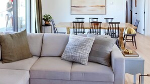 A modern living room with a sofa, a dining table with chairs, a large abstract painting, and a sliding glass door leading to an outdoor area.