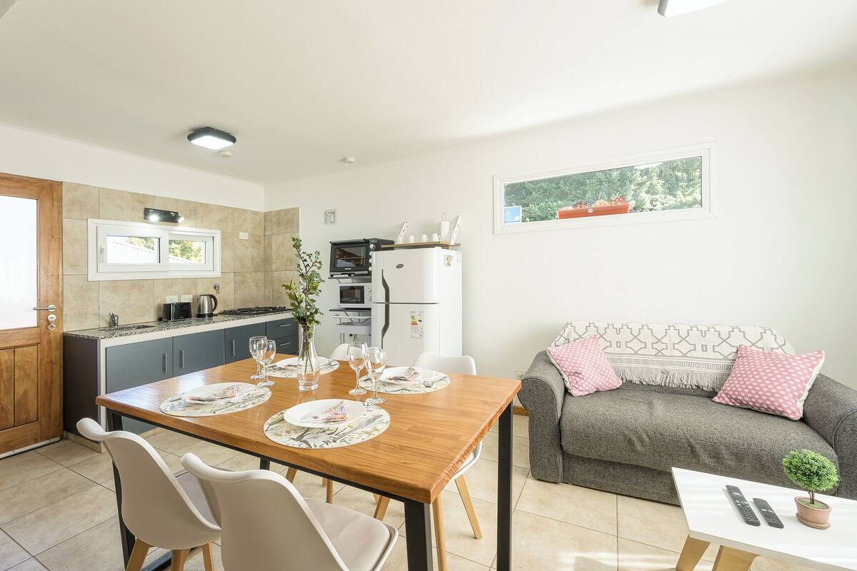 Une cuisine compacte dotée d’appareils électroménagers modernes, un coin repas avec une table et des chaises en bois, et un espace de vie chaleureux avec un canapé et des coussins.