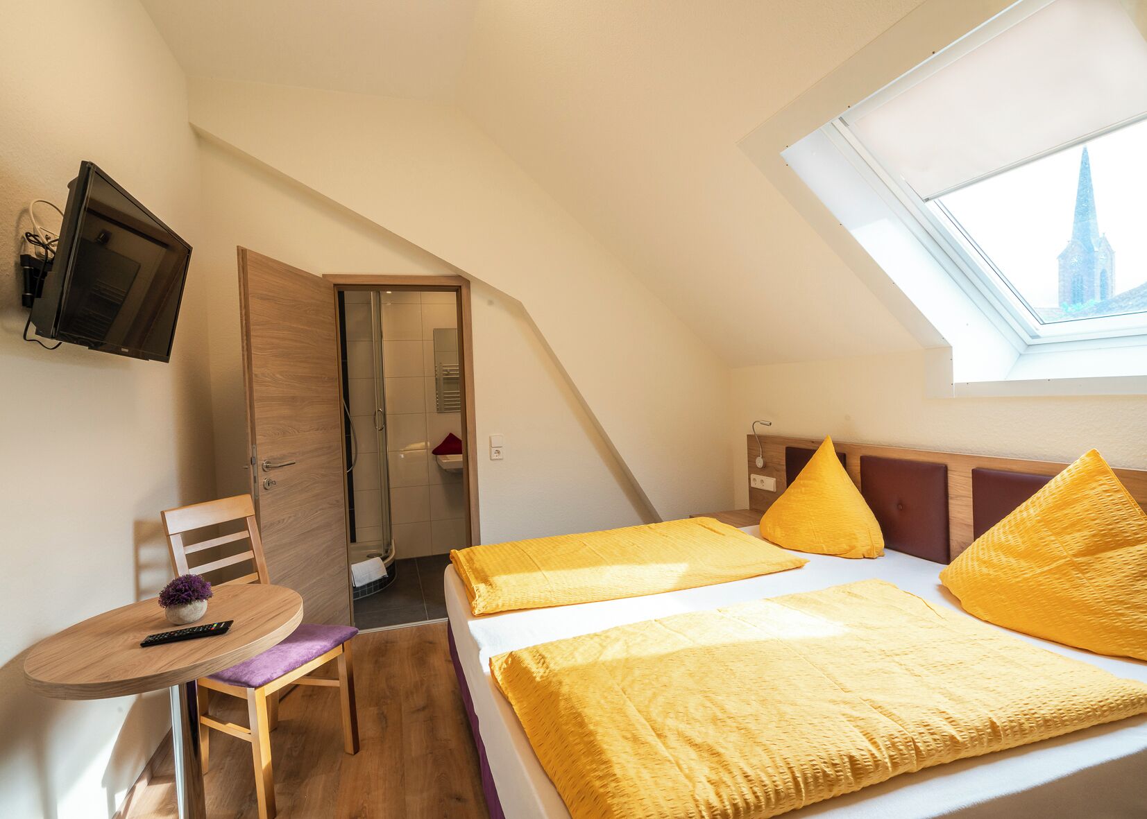 Ein kleines, modernes Schlafzimmer mit schrägem Dach, einem Bett mit gelber Bettwäsche, einem an der Wand montierten Fernseher und einem Fenster mit Blick auf ein Gebäude.