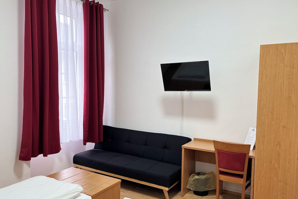 A small, modern room with a black sofa, a wooden desk, a TV mounted on the wall, and a window with red curtains.