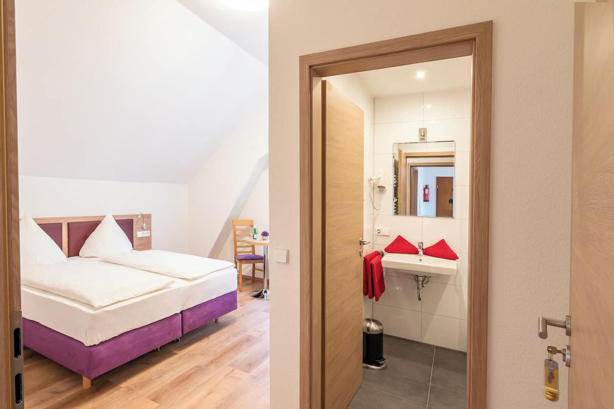 A bedroom with a bed, a chair, and a small table. There is a bathroom with a sink and a mirror. The room has wooden flooring and a sloped ceiling.