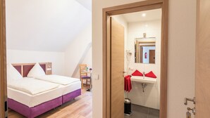 Un dormitorio con una cama, una silla y una mesita. Hay un baño con lavamanos y espejo. El cuarto tiene piso de madera y techo inclinado.