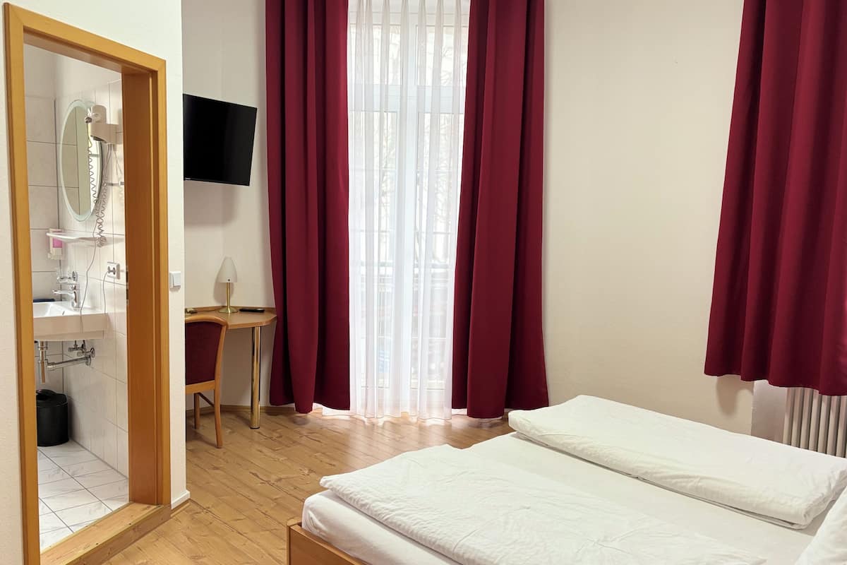 A hotel room with a wooden floor, a bed with white bedding, a desk with a lamp, a TV mounted on the wall, and a window with red curtains.