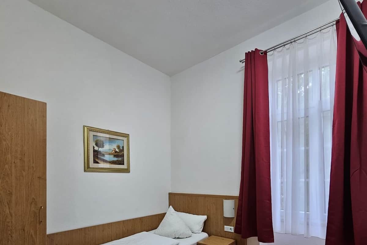 A single bed with a wooden headboard, a bedside table, a small cabinet, a chair, a desk with a remote control, and a window with red curtains.