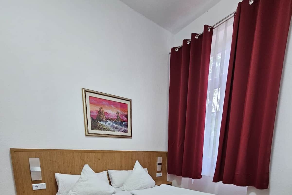 A double bed with a wooden headboard, white bedding, a nightstand, a painting on the wall, and red curtains on a window.
