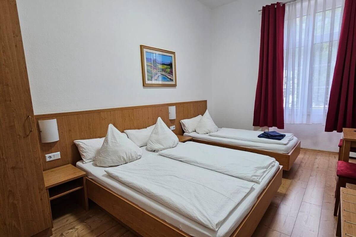 A hotel room with two beds, wooden flooring, a painting on the wall, and a window with red curtains.