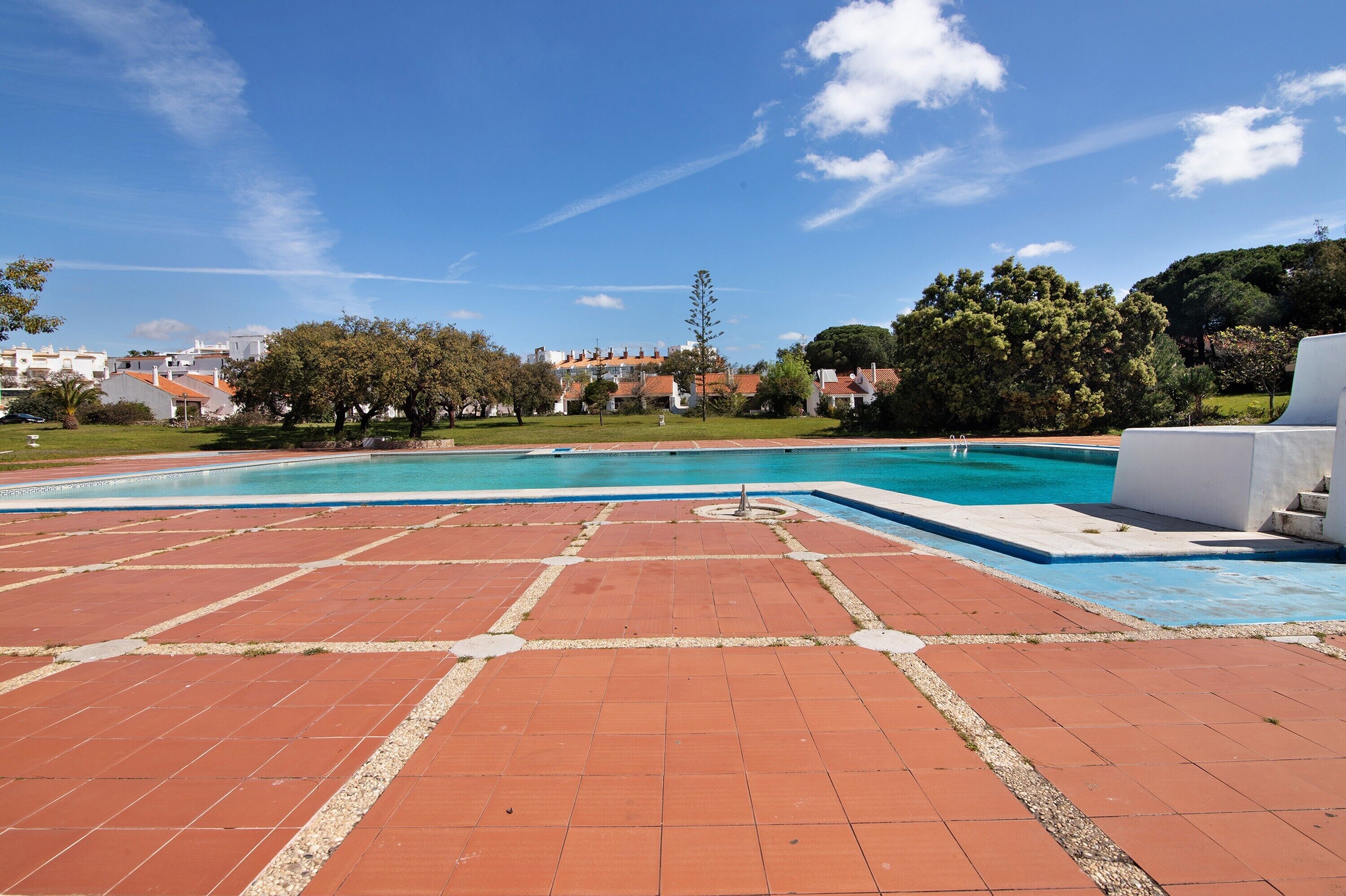 Una piscina con borde de baldosas, rodeada de vegetación y edificios al fondo.