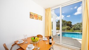 Un coin repas avec une table dressée pour deux personnes, un panier de fruits, du vin et une vue sur une piscine.