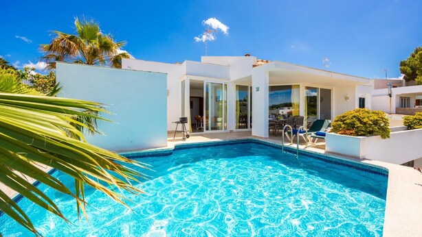 Un espace moderne aménagé au bord de la piscine, avec une piscine à l’eau d’un bleu limpide, un coin salon abrité et un bâtiment blanc doté de portes vitrées.