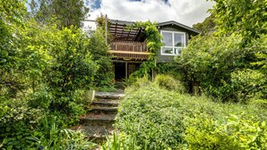 Ein Haus mit Holzterrasse und überdachter Veranda, umgeben von üppigem Grün und einem Kiesweg, der zum Eingang führt.