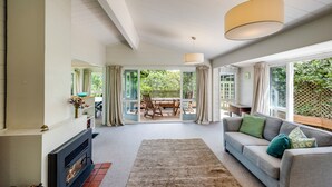 Ein geräumiges Wohnzimmer mit Kamin, einer grauen Couch mit grünen und türkisen Kissen, einem großen Teppich und Blick auf eine Terrasse mit Holztisch und Stühlen.